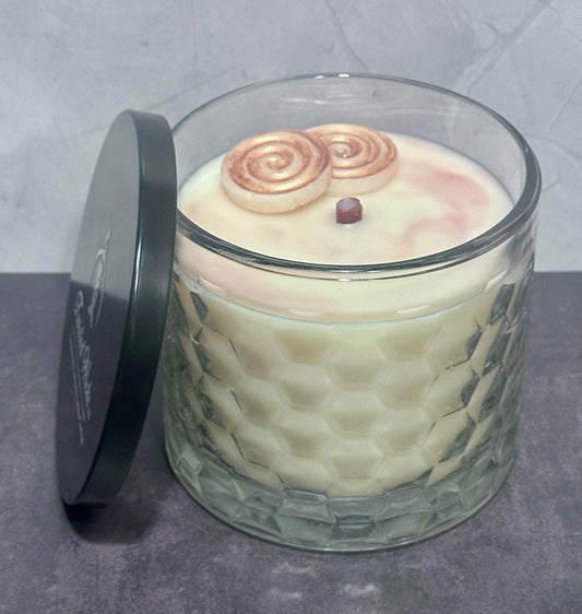 Sticky Situation 12oz artisan candle in a textured honeycomb pattern container, filled with creamy off-white wax and topped with two cinnamon roll wax embeds. A black metal lid with white script branding leans against the left side of the glass jar on a gray surface.