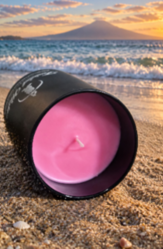 Eruption 7oz artisan candle in a matte black glass tumbler lying on its side in the sand to show the pink wax, with ocean waves and a mountain at sunset behind it
