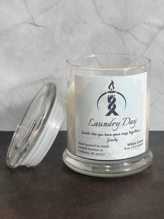 Laundry Day 8oz artisan candle in a glass jar candle with its lid leaning beside it on a dark tabletop. The candle has a white label reading 'Laundry Day' with a the Twisted Wicks logo