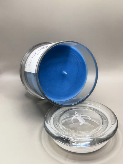 A clear glass jar candle with blue wax, shown tipped on its side with the glass lid placed in front on a light gray background.