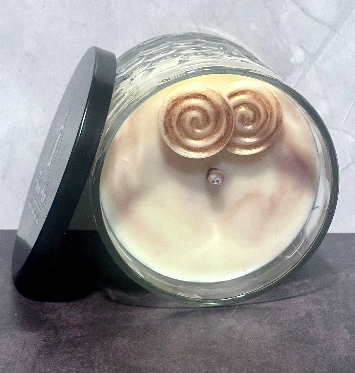 Sticky Situation 12oz artisan candle lying on its side, light beige wax topped with two cinnamon roll wax embeds and dusted with bronze mica