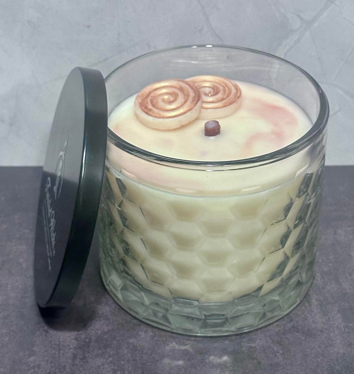 Sticky Situation 12oz artisan candle in a textured honeycomb pattern container, filled with creamy off-white wax and topped with two cinnamon roll wax embeds. A black metal lid with white script branding leans against the left side of the glass jar on a gray surface.